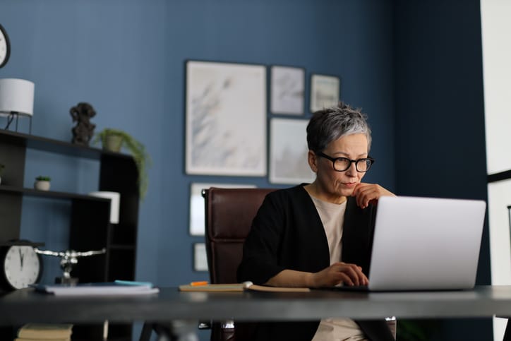 Photo shows a mature businesswoman sitting confidently at her modern office desk, focused on her laptop/Getty Images