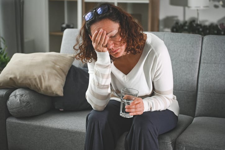 Woman sitting on couch with head in hand, holding glass of water