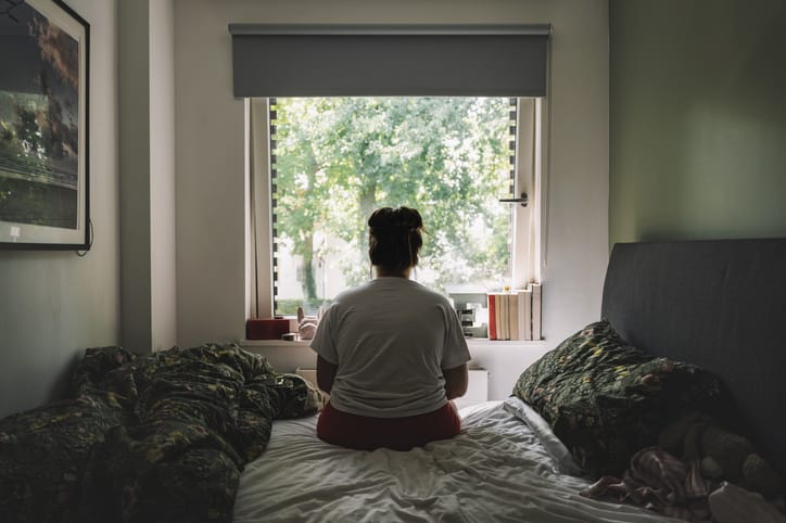 Photo shows a woman sitting on her bed and looking out the window/Getty Images